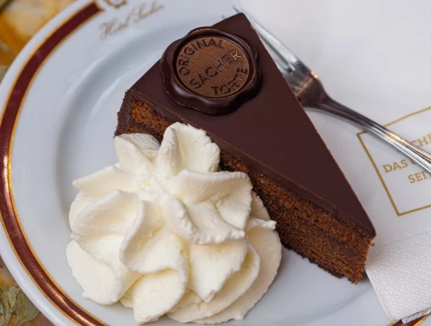Ein Stück Original Sacher-Torte mit Sahne und Gabel auf edlem Porzellanteller serviert.