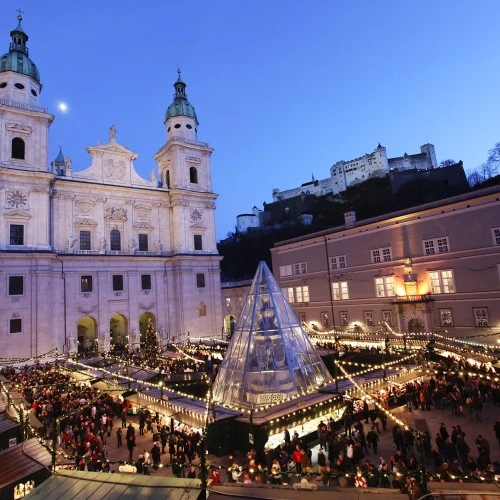 Belebter Weihnachtsmarkt vor dem Salzburger Dom in festlicher Beleuchtung, im Hintergrund die Festung Hohensalzburg.