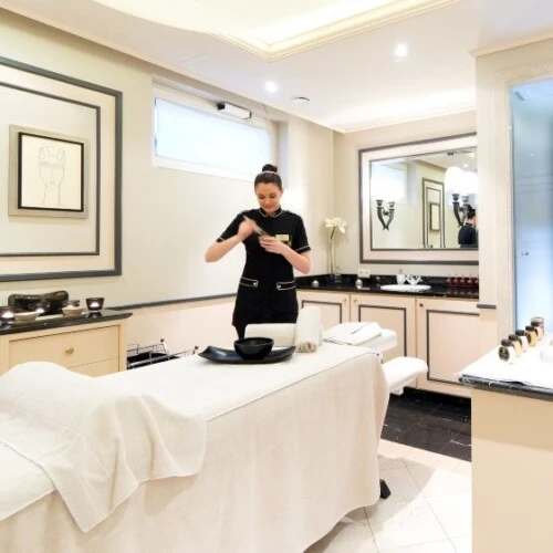 Heller, eleganter Behandlungsraum eines Spas mit Massageliege, auf der eine weiße Decke liegt; eine Mitarbeiterin in schwarzer Uniform bereitet eine Behandlung vor, umgeben von dezentem Dekor und luxuriösen Pflegeprodukten.