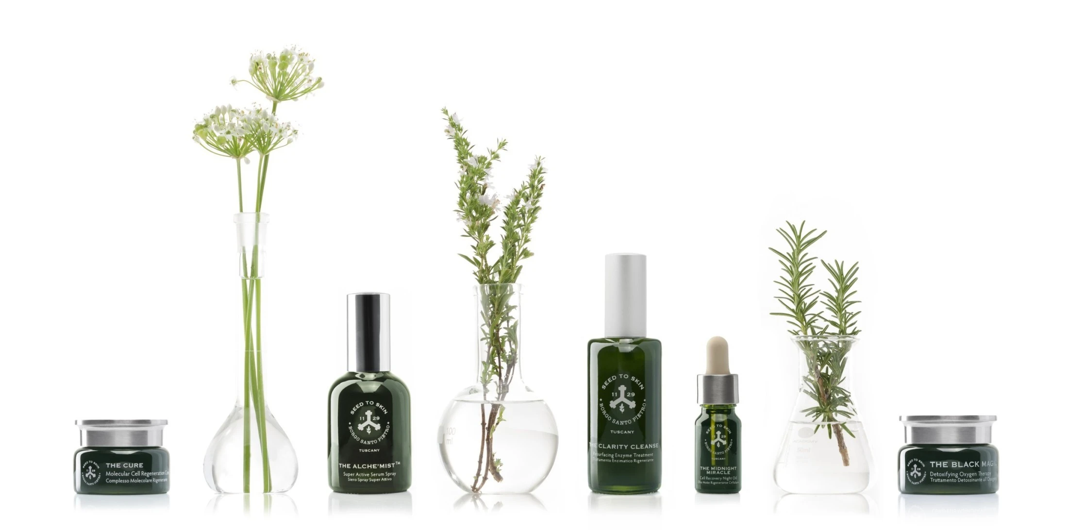 Produktlinie von „Seed to Skin“ in grünen Glasbehältern, arrangiert zwischen Laborfläschchen mit frischen Kräutern und Blüten auf weißem Hintergrund.