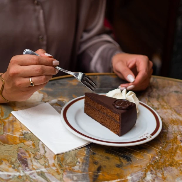 Ein Stück Sachertorte mit Sahne wird auf einem eleganten Teller auf einem kunstvoll gemaserten Marmortisch serviert, während eine Person mit einer Gabel ein Stück davon nimmt.