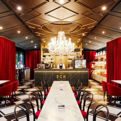 Ein elegantes Café mit schwarz-weiß gemustertem Boden, Marmortischen und roten Samtstühlen, im Zentrum eine goldene Bar mit großer Kristallleuchter darüber und roten Samtvorhängen an den Wänden.