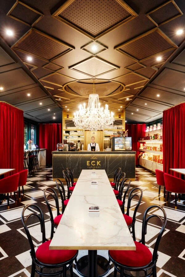 Ein elegantes Café mit schwarz-weiß gemustertem Boden, Marmortischen und roten Samtstühlen, im Zentrum eine goldene Bar mit großer Kristallleuchter darüber und roten Samtvorhängen an den Wänden.