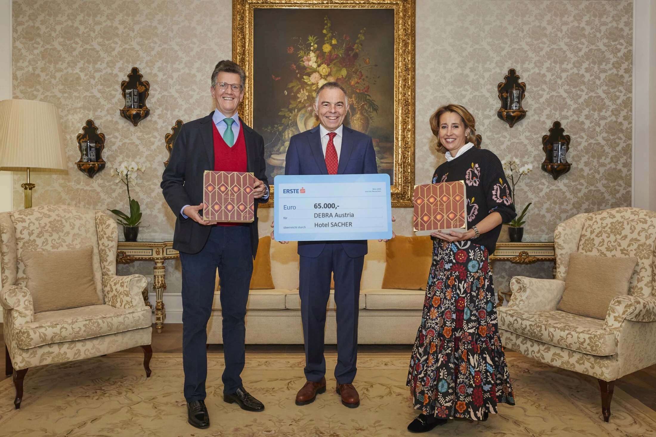 Drei Personen stehen vor einer eleganten Wand mit floralem Muster, flankiert von zwei Sesseln. Der Mann in der Mitte hält einen großen Spendenscheck von 65.000 Euro für DEBRA Austria vom Hotel Sacher, während die anderen beiden jeweils eine edle Sacher-Box mit rot-goldenem Muster präsentieren.