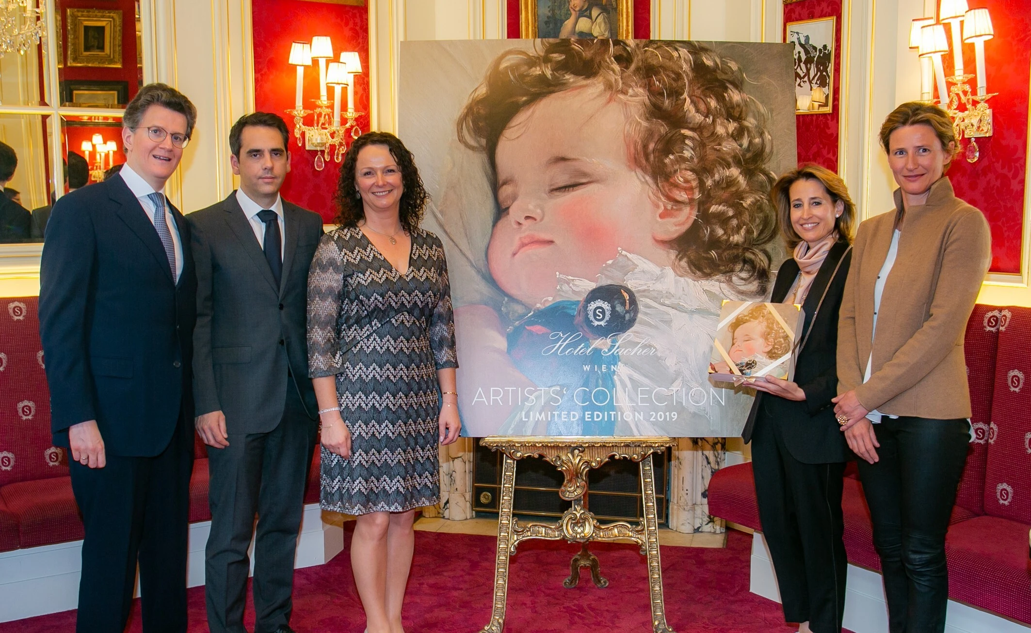 Sechs Personen stehen vor einem großen Kunstwerk eines schlafenden Kindes, das die Sacher Artists' Collection 2019 präsentiert. Eine Frau hält eine kleinere Box mit demselben Motiv in den Händen. Die Umgebung ist elegant mit rotem Interieur und goldenen Akzenten gestaltet.
