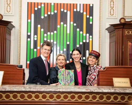 Vier Personen stehen lächelnd hinter einer Rezeption vor einem bunten Kunstwerk mit weißen Punkten und farbigen Streifen.