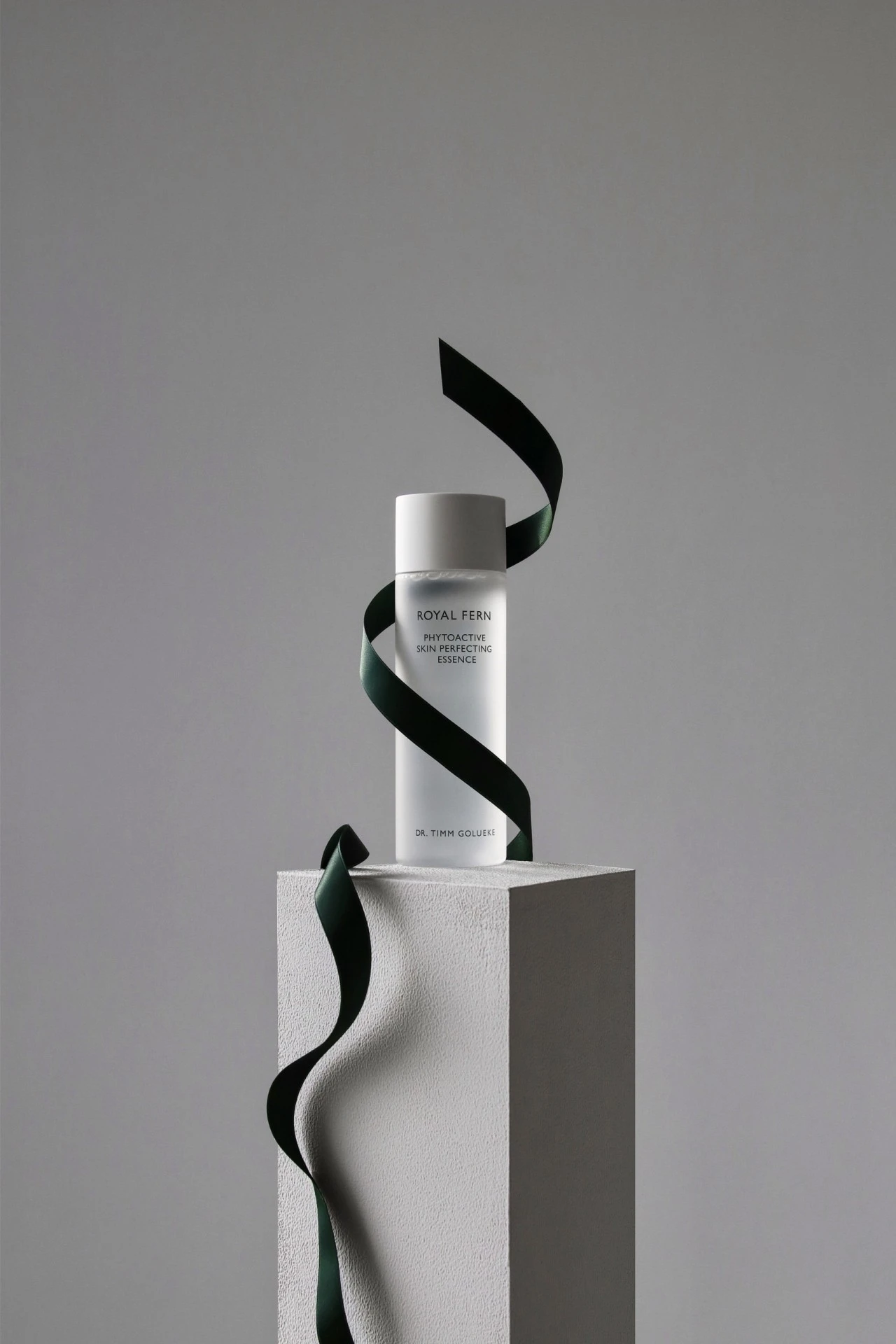 Eine Flasche Royal Fern Phytoactive Skin Perfecting Essence steht auf einem weißen Podest, elegant umschlungen von einem grünen Satinband.