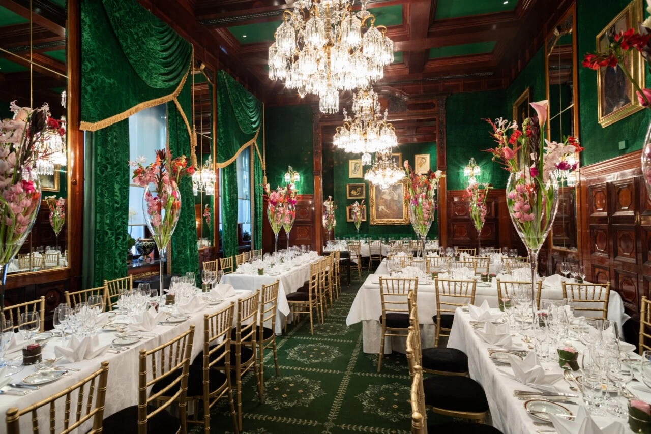 Prunkvoll dekorierten Festsaal mit einer üppigen und eleganten Atmosphäre. Der Raum ist geprägt von dunkelgrünen Wänden und Vorhängen, glänzenden Holzelementen und großen Kristalllüstern, die von der Decke hängen und warmes Licht spenden