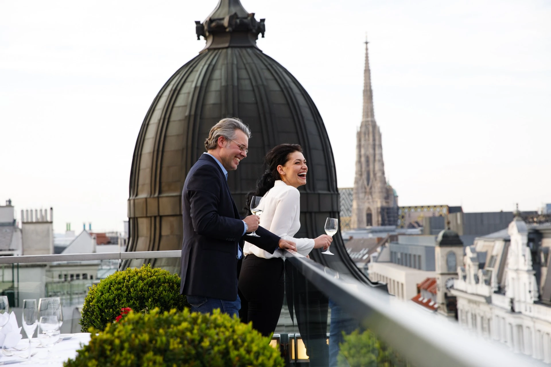 Lachendes Paar auf einer Dachterrasse. Sie trinken Weißwein und genießen die Aussicht über Wien. Im Hintergrund sind eine große Kuppel und der Stephansdom zu sehen. Die Stimmung ist fröhlich und entspannt.