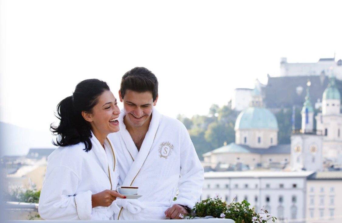 Ein Paar in weißen Bademänteln mit Sacher-Logo auf stehen auf dem Balkon. Beide lachen und genießen gemeinsam eine Tasse Kaffee. Im Hintergrund ist die Silhouette von Salzburg mit der Festung Hohensalzburg und den Kuppeln des Doms zu sehen.