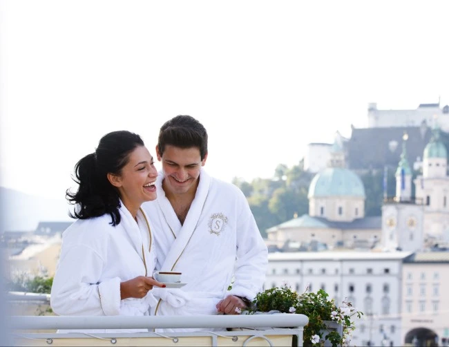 Ein Paar in weißen Bademänteln mit Sacher-Logo auf stehen auf dem Balkon. Beide lachen und genießen gemeinsam eine Tasse Kaffee. Im Hintergrund ist die Silhouette von Salzburg mit der Festung Hohensalzburg und den Kuppeln des Doms zu sehen.