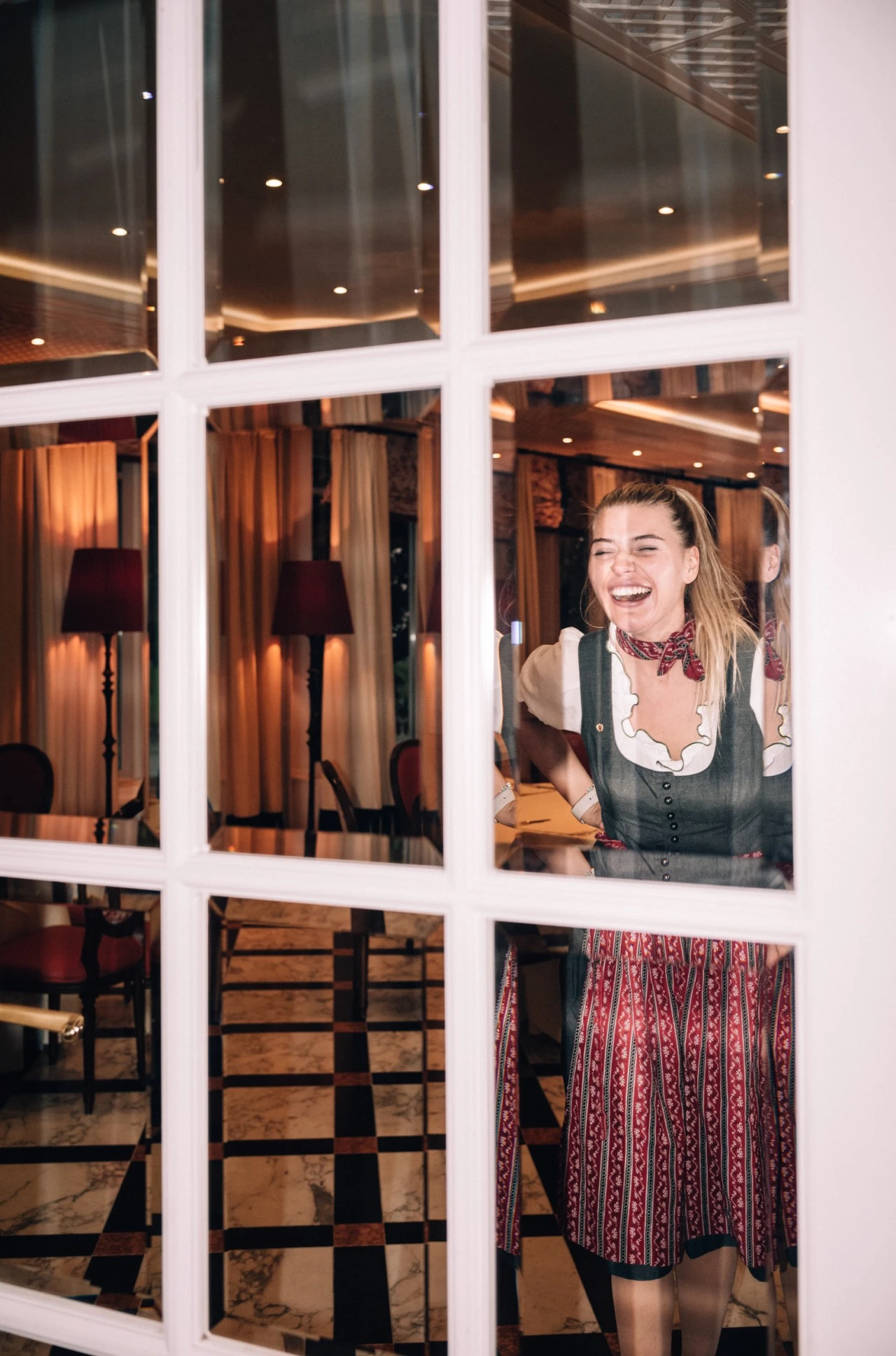 Eine junge Frau in traditionellem Dirndl steht lachend hinter einer gläsernen Tür. Sie trägt ein dunkelgrünes Mieder mit weißer Bluse und ein rot-gemustertes Rockteil.