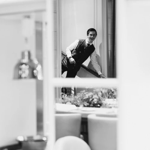 Ein Kellner in traditioneller Uniform wird in einem eleganten Schwarzweiß-Foto eingefangen, wie er mit einem freundlichen Lächeln eine Treppe hinuntersteigt.