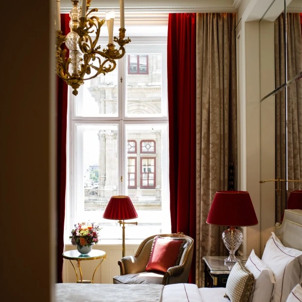 Elegantes Hotelzimmer mit Blick auf ein historisches Gebäude durch hohe Fenster. Das Interieur ist in warmen Tönen gehalten: ein einladendes Bett mit dekorativen Kissen, klassische Nachttischlampen mit roten Schirmen, ein goldener Sessel mit rotem Polster sowie ein Blumenarrangement auf einem Beistelltisch. Ein Kronleuchter rundet das luxuriöse Ambiente ab.