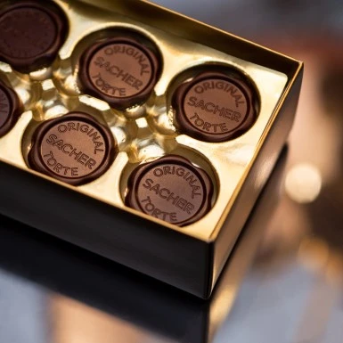 Goldene Pralinenschachtel mit sechs runden Schokoladenstücken zu sehen. Jedes Stück trägt das geprägte Siegel „ORIGINAL SACHER TORTE“, das für die weltberühmte Wiener Spezialität steht.
