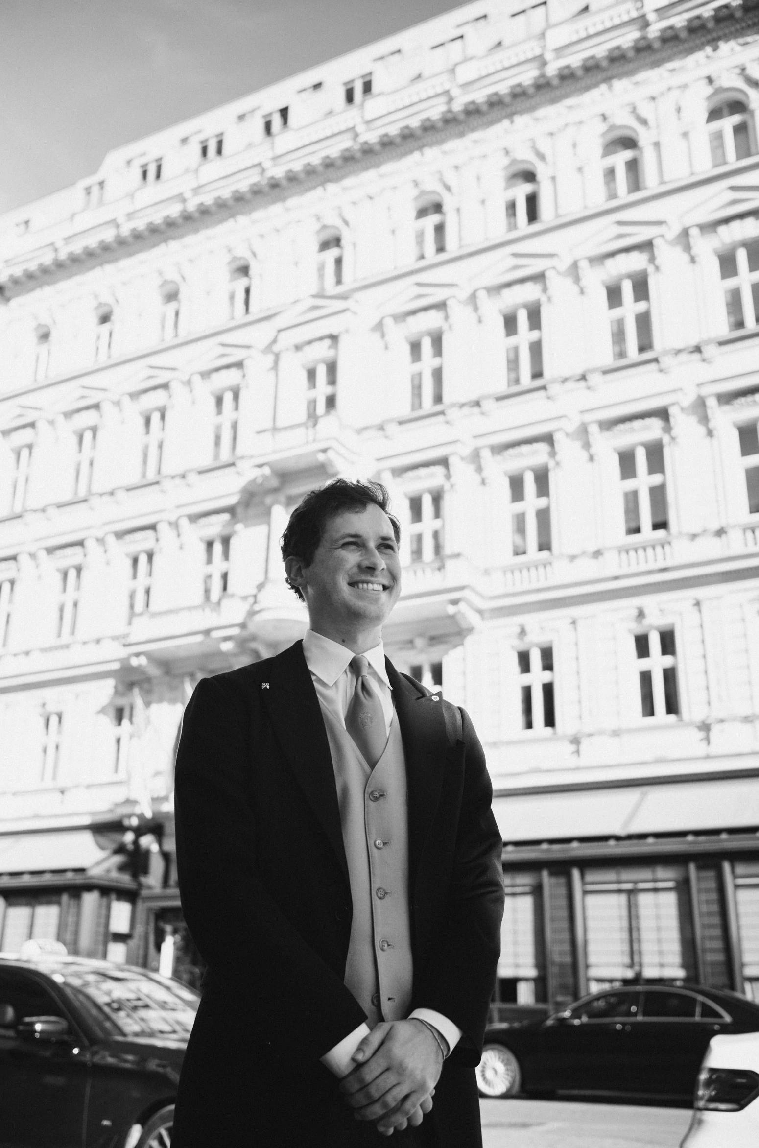 Im Vordergrund steht ein lächelnder Mitarbeiter im eleganten Frack mit Weste und Krawatte – klassisch und stilvoll. Im Hintergrund ist die Fassade des Hotel Sacher Wien zu sehen, mit ihren prunkvollen Fensterrahmen und historischen Architekturelementen. Eine schwarze Limousine verstärkt das exklusive Ambiente vor dem Eingang.