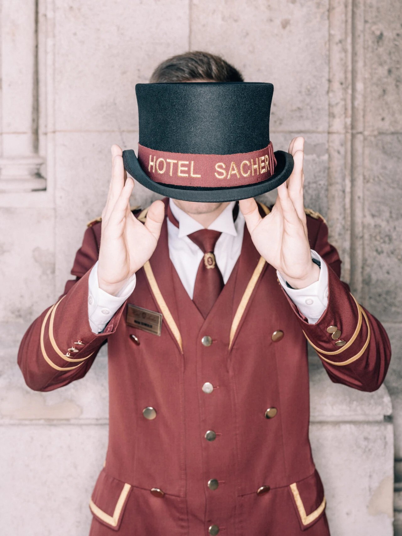 Ein Portier in der klassischen, burgunderfarbenen Uniform des Hotel Sacher hält einen Zylinder mit goldener Aufschrift „HOTEL SACHER“ vor sein Gesicht.