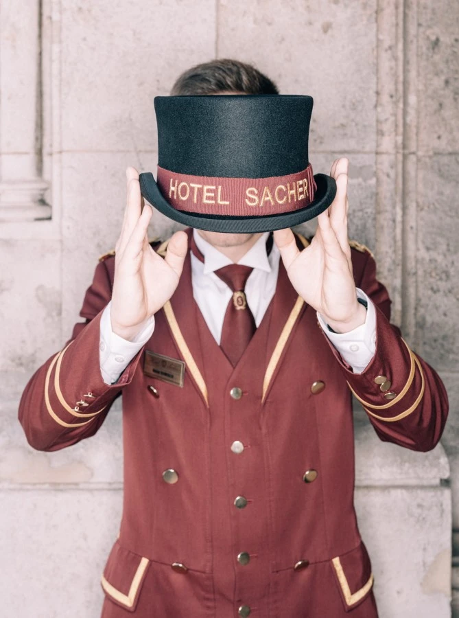 Hotel Sacher Salzburg, Concierge Ein Portier in der klassischen, burgunderfarbenen Uniform des Hotel Sacher hält einen Zylinder mit goldener Aufschrift „HOTEL SACHER“ vor sein Gesicht.