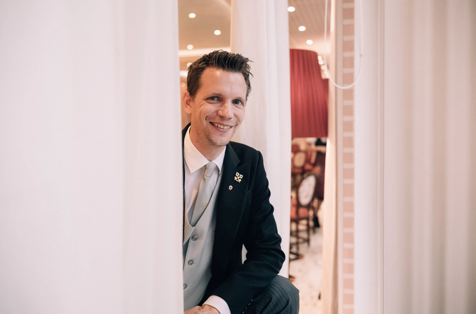 Ein elegant gekleideter Concierge mit einem freundlichen Lächeln sitzt zwischen weißen Vorhängen.