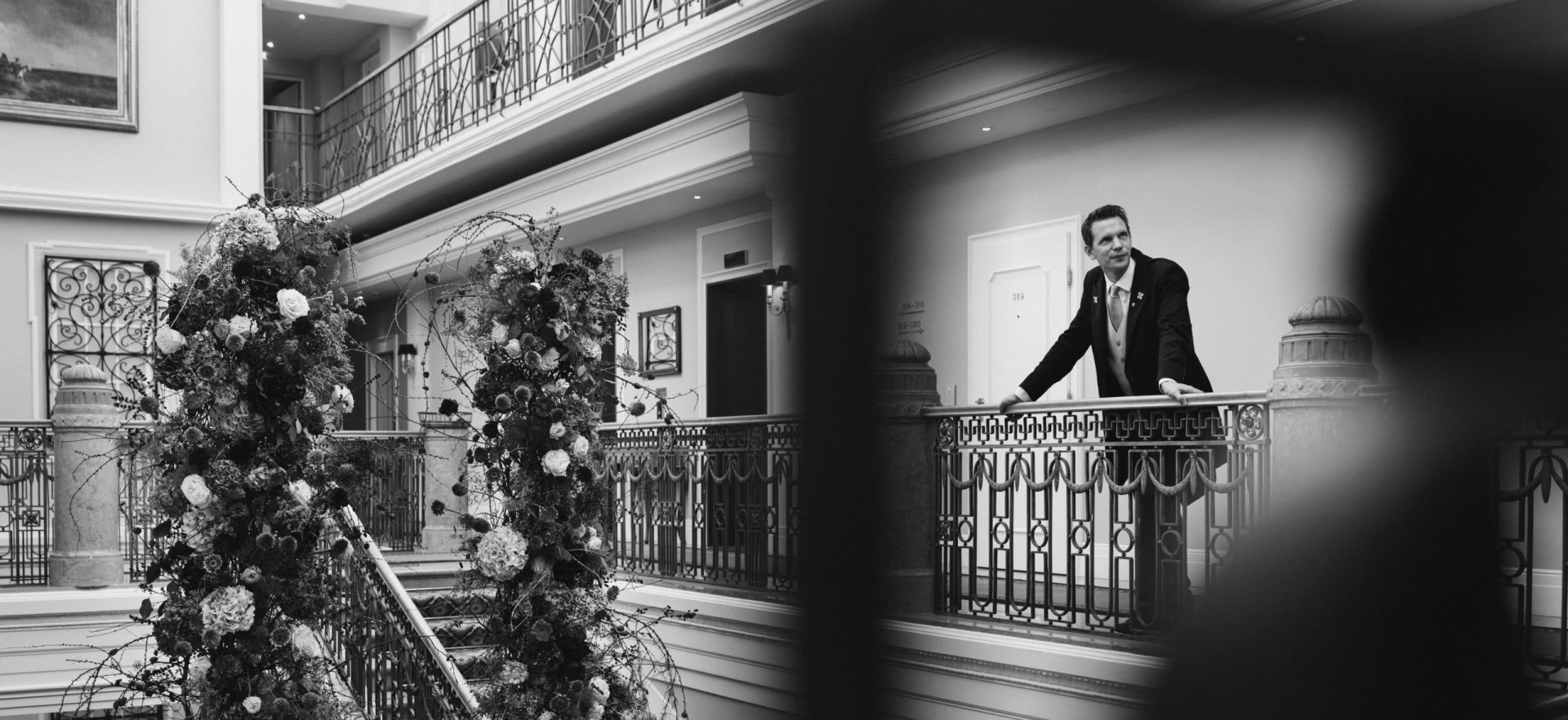 Elegant gekleideter Concierge lehnt am kunstvoll verzierten Geländer im oberen Stockwerk eines Hotels. Im Vordergrund florale Dekoration an einem Treppengeländer. Die Aufnahme ist in Schwarz-Weiß