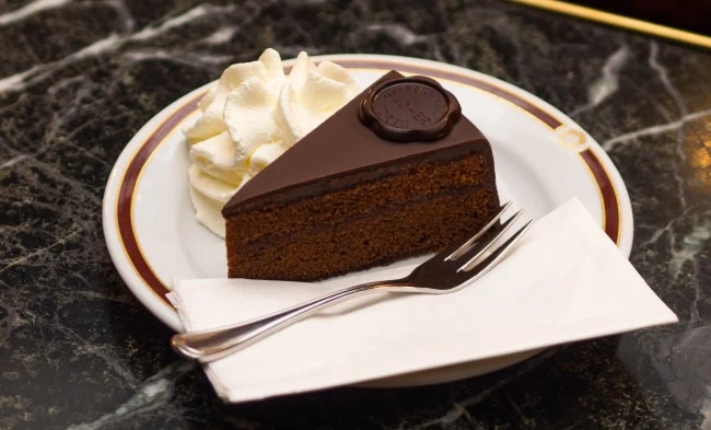 Ein Stück Original Sacher-Torte mit glänzender Schokoglasur und der typischen Schokoladenplakette, daneben eine großzügige Portion Schlagobers. Serviert auf feinem Porzellanteller mit goldbraunem Rand, angerichtet auf einem eleganten, dunklen Marmortisch.