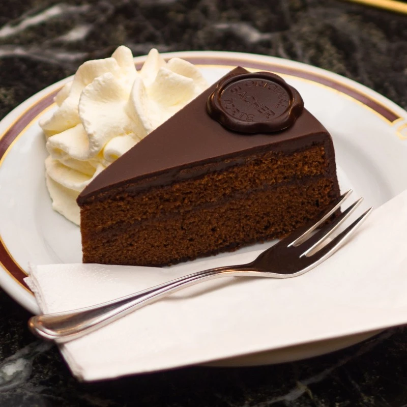 Ein Stück Original Sacher-Torte mit glänzender Schokoglasur und der typischen Schokoladenplakette, daneben eine großzügige Portion Schlagobers. Serviert auf feinem Porzellanteller mit goldbraunem Rand, angerichtet auf einem eleganten, dunklen Marmortisch.