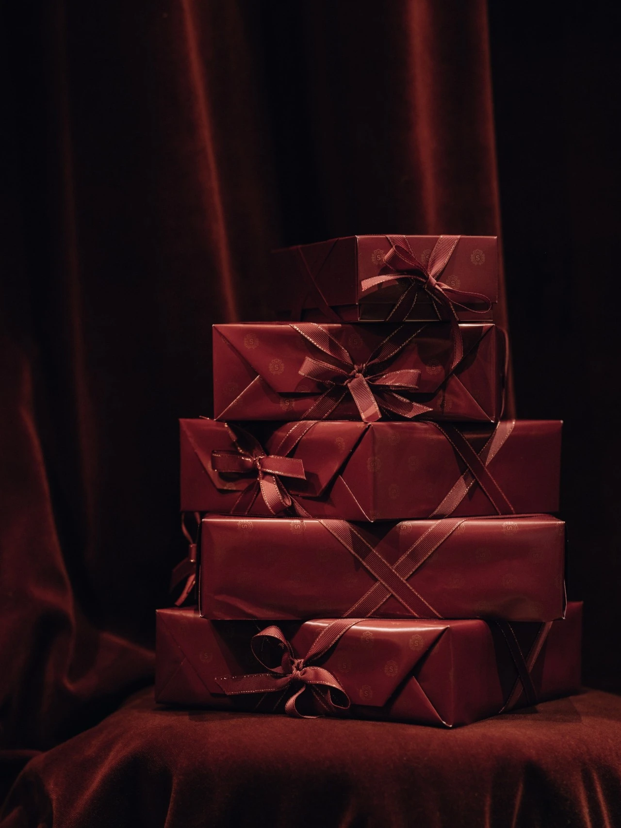 Ein Stapel sorgfältig verpackter Geschenkboxen in tiefem Sacher-Bordeaux, veredelt mit seidigen Schleifen. Das Papier trägt dezent das geprägte Sacher-Logo und wirkt in Kombination mit dem dunklen Samt-Hintergrund besonders edel und geheimnisvoll.