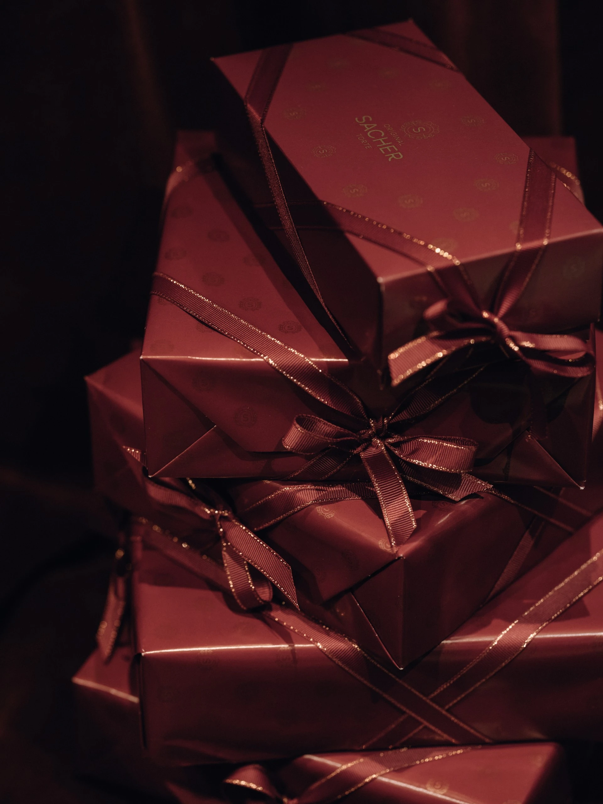 Ein Stapel sorgfältig verpackter Geschenkboxen in tiefem Sacher-Bordeaux, veredelt mit seidigen Schleifen. Das Papier trägt dezent das geprägte Sacher-Logo und wirkt in Kombination mit dem dunklen Samt-Hintergrund besonders edel und geheimnisvoll.