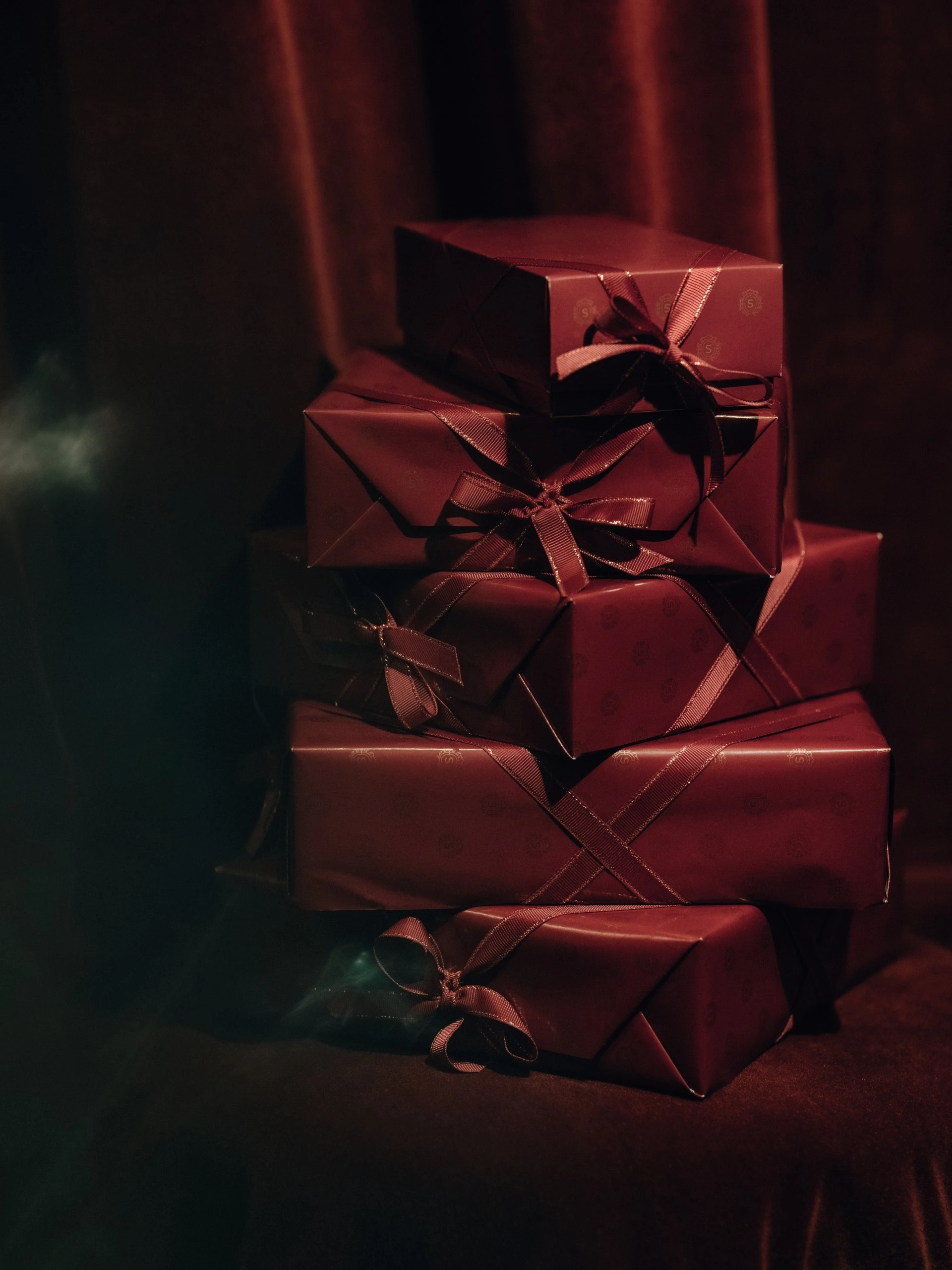 Ein Stapel sorgfältig verpackter Geschenkboxen in tiefem Sacher-Bordeaux, veredelt mit seidigen Schleifen. Das Papier trägt dezent das geprägte Sacher-Logo und wirkt in Kombination mit dem dunklen Samt-Hintergrund besonders edel und geheimnisvoll.