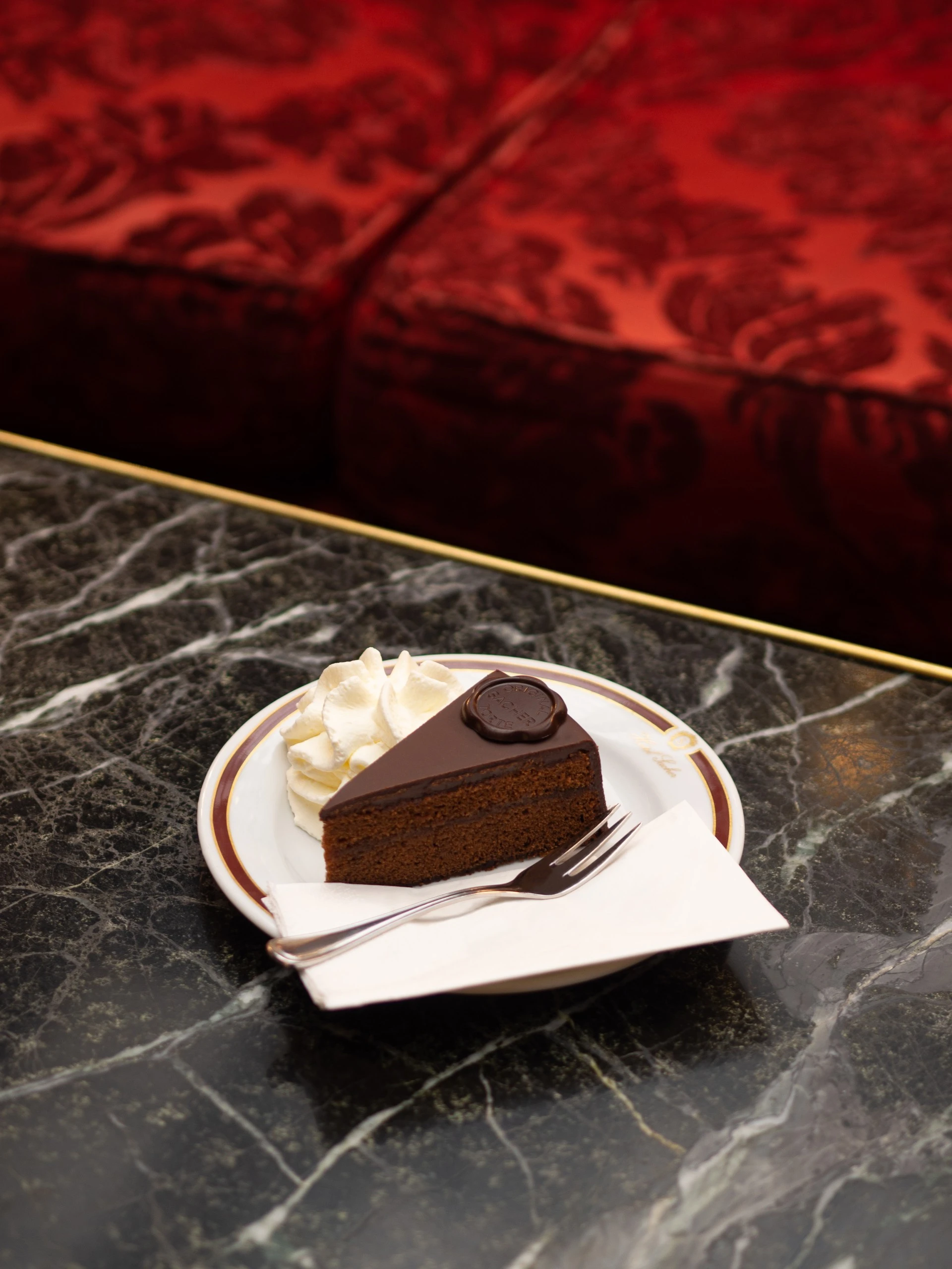 Ein Stück Original Sacher-Torte mit typischer Schokoladenplakette und einem Klecks Schlagobers, serviert auf Porzellan mit Goldrand. Der Teller steht auf einem dunklen Marmortisch, im Hintergrund ein samtiges, rot gemustertes Sofa – elegant und zeitlos.