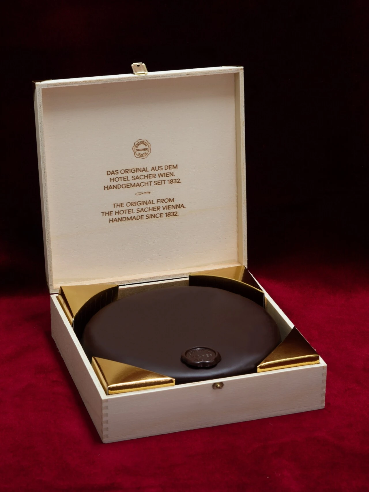 Eine ganze Original Sacher-Torte liegt stilvoll verpackt in einer eleganten Holzschatulle mit goldenen Ecken. Auf dem Deckel der Box der Hinweis auf ihre handgemachte Herkunft aus dem Hotel Sacher Wien seit 1832.