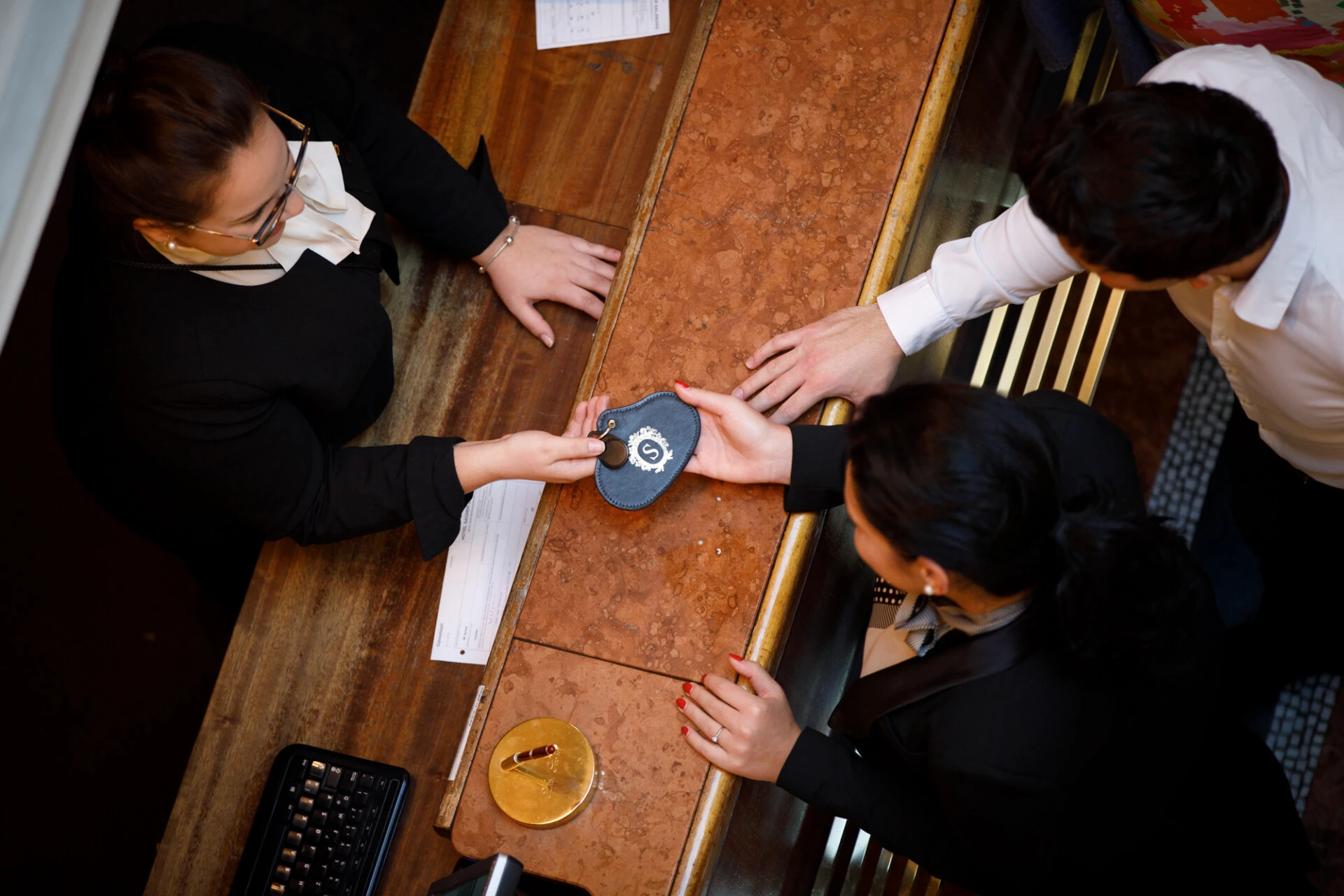 Ein Moment an der Rezeption – zwei Gäste nehmen mit einem Lächeln den traditionellen Zimmerschlüssel mit Lederanhänger entgegen. Die Szene wirkt herzlich und professionell, mit warmem Licht, eleganter Kleidung und einem Hauch klassischer Gastfreundschaft.