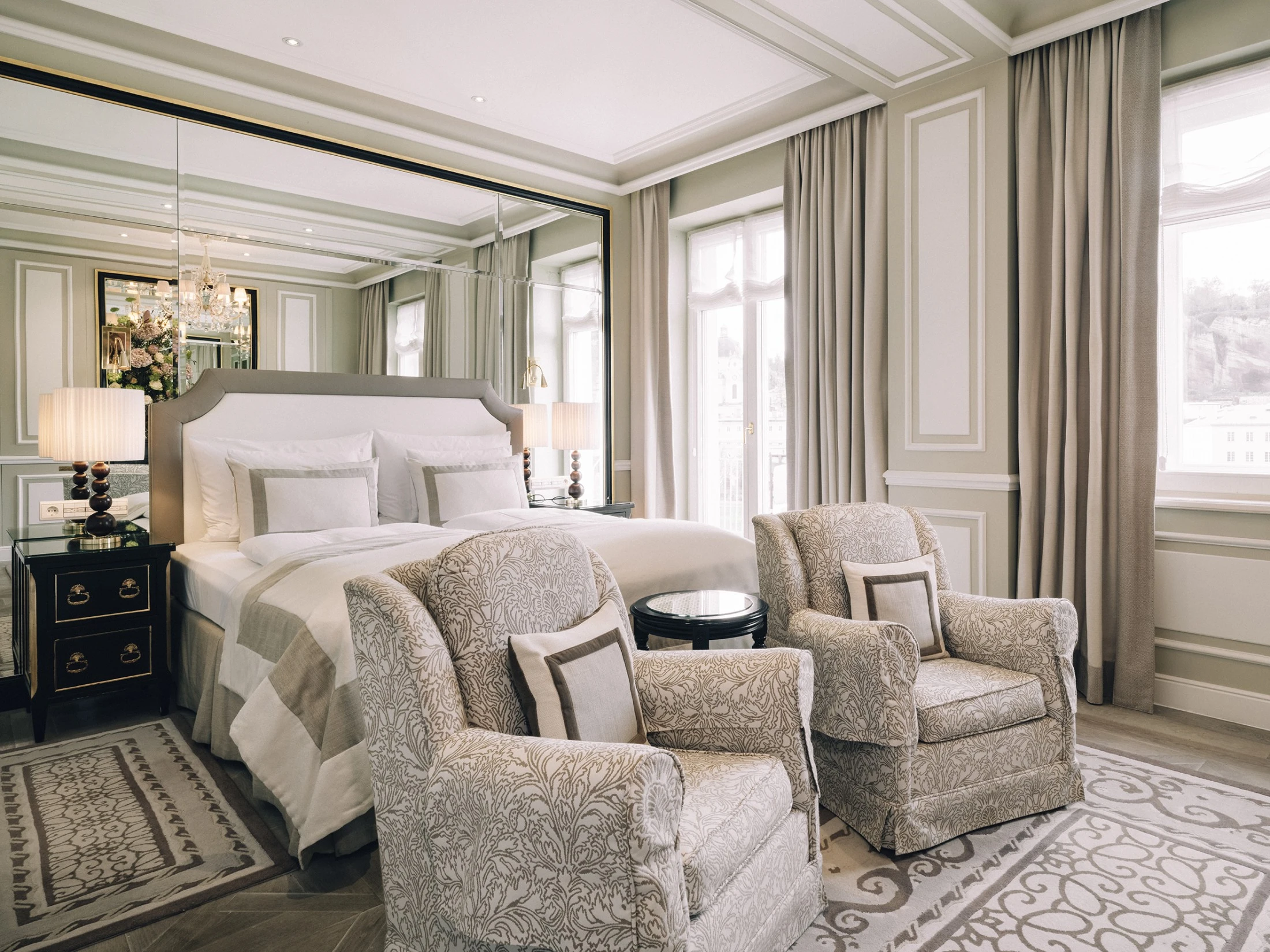 Elegantes Schlafzimmer mit einem großen Doppelbett, zwei gemütlichen Sesseln mit floraler Musterung, seitlichen Nachttischen mit klassischen Lampen und bodentiefen Fenstern.