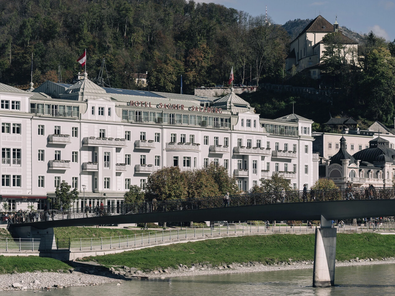 Hotel Sacher Salzburg am Tag. Im Vordergrund sieht man eine Brücke, die über die Salzach führt.