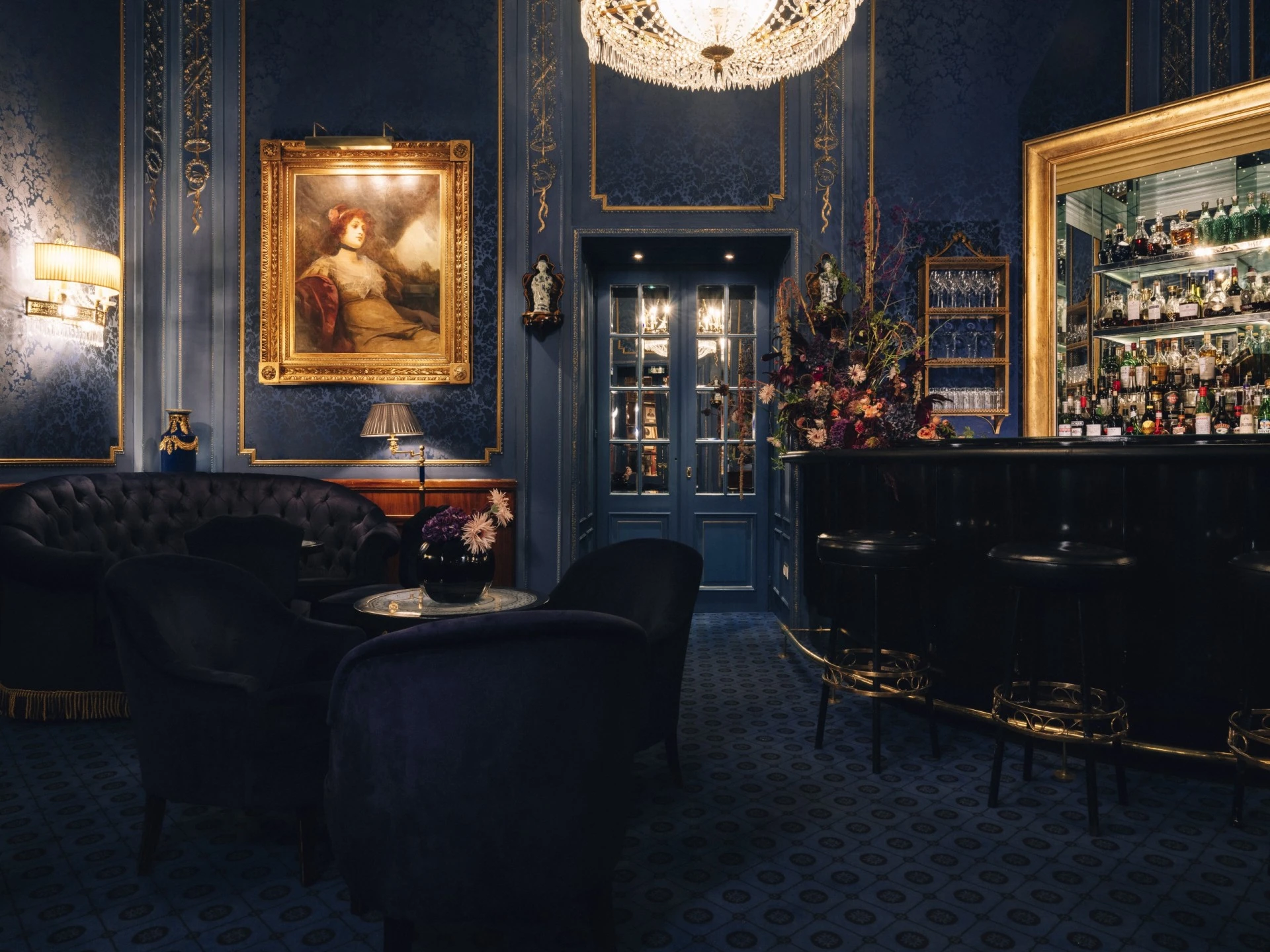 Stilvolle Bar-Lounge mit tiefblauer, opulenter Einrichtung und goldenen Akzenten. Die Wände sind mit gemustertem Stoff bespannt, ein prächtiger Kronleuchter spendet warmes Licht. Im Zentrum der Szene steht die elegante Bar mit verspiegeltem Regal voller hochkarätiger Spirituosen.
