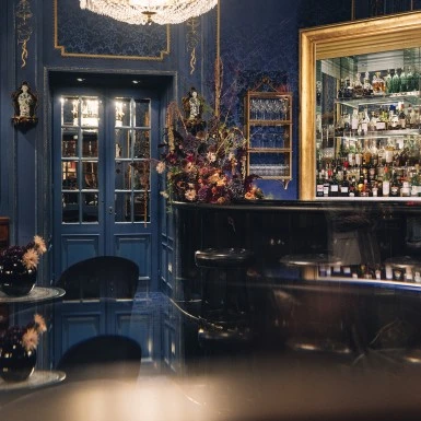 Ein eleganter Barbereich in sattem Königsblau, durchzogen von goldenen Akzenten. Die Spiegelwand hinter der Bar reflektiert die sorgfältig arrangierten Flaschen und verleiht dem Raum zusätzliche Tiefe. Die florale Komposition am Tresen setzt einen farbintensiven Kontrapunkt.