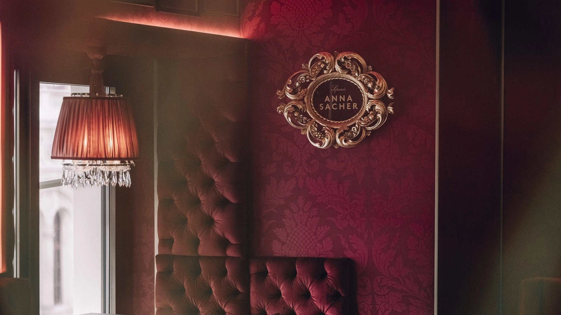 Im Zentrum des Bildes hängt ein kunstvoll gerahmtes Emblem mit der Aufschrift „Salon Anna Sacher“. Links fällt Tageslicht durch ein Fenster, darunter hängt eine klassische Lampe mit Kristallbehang, die warmes Licht spendet.