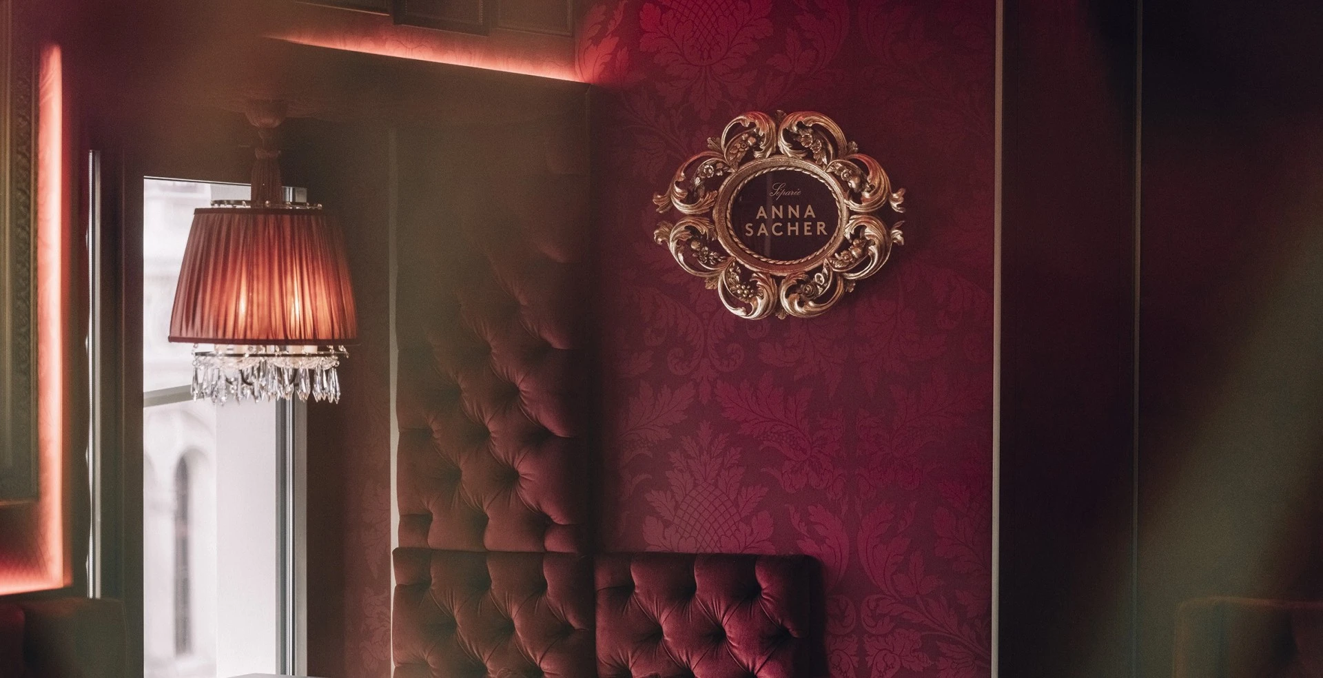 Im Zentrum des Bildes hängt ein kunstvoll gerahmtes Emblem mit der Aufschrift „Salon Anna Sacher“. Links fällt Tageslicht durch ein Fenster, darunter hängt eine klassische Lampe mit Kristallbehang, die warmes Licht spendet.