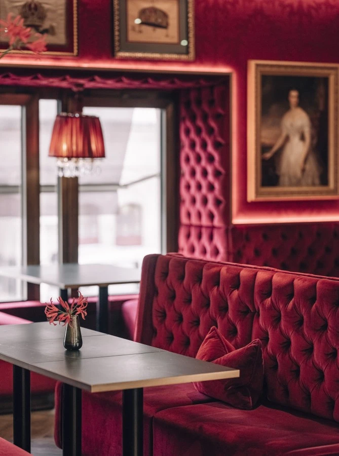 Hotel Sacher Wien, Café Bel Étage Ein stilvoller Sitzbereich mit gepolsterten, samtroten Bänken und schwarzen Tischen. Die Wände sind mit klassischen Gemälden und historischen Bildern geschmückt.