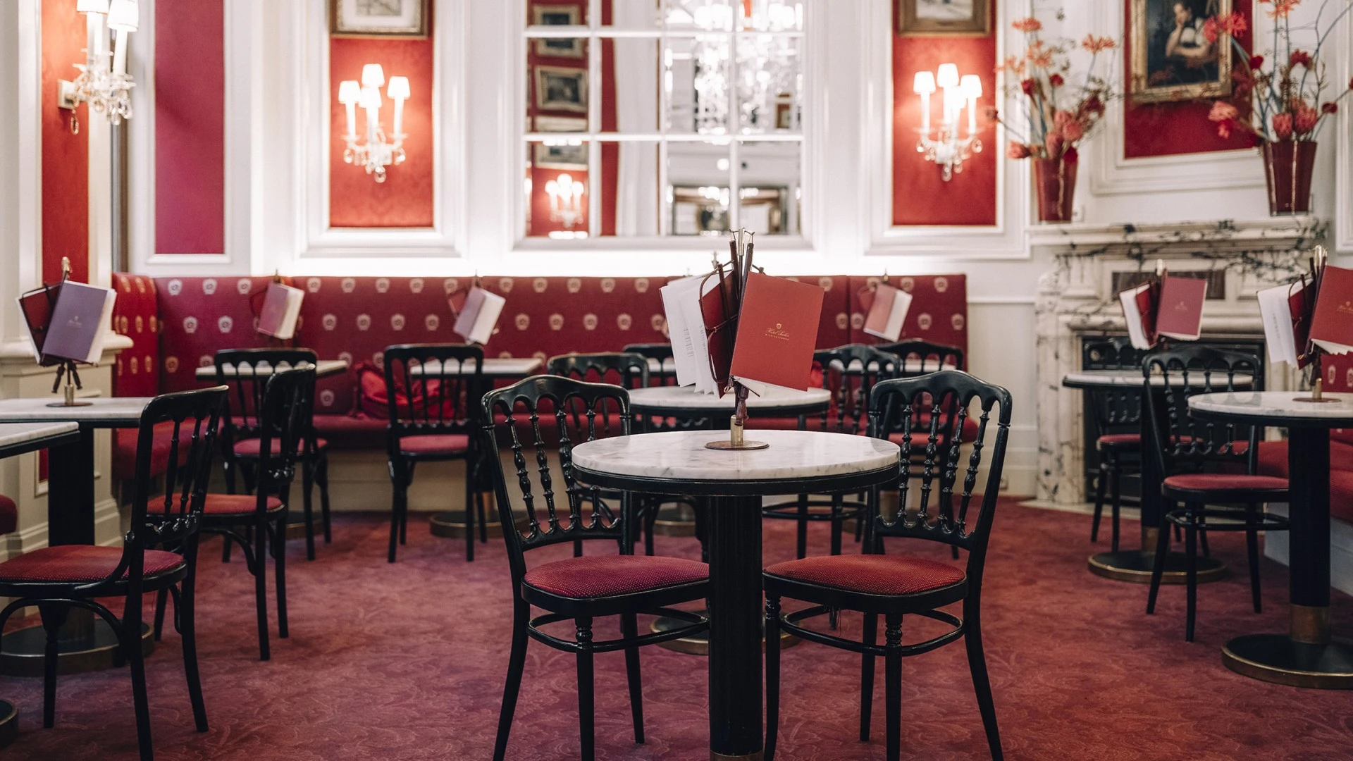 Ein stilvoll eingerichtetes Café mit eleganten Marmortischen und rot gepolsterten Stühlen. Die Wände sind mit historischen Bildern und Gemälden geschmückt, eingerahmt von roten Stoffpaneelen. Kristalllüster und Wandleuchten sorgen für ein warmes Licht.