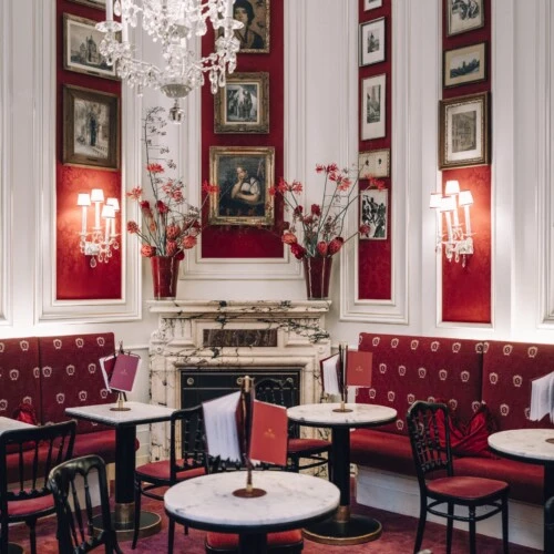 Ein stilvoll eingerichtetes Café mit eleganten Marmortischen und rot gepolsterten Stühlen. Die Wände sind mit historischen Bildern und Gemälden geschmückt, eingerahmt von roten Stoffpaneelen. Kristalllüster und Wandleuchten sorgen für ein warmes Licht.
