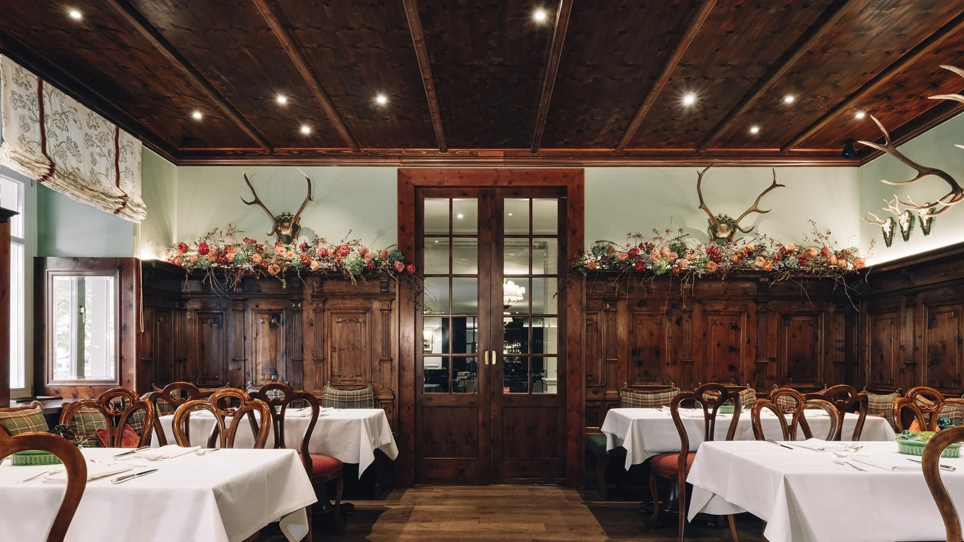 Rustikales Restaurant (Zirbelzimmer) mit dunklem Holzinterieur, weißen Tischdecken, Hirschgeweihen an der Wand und karierten Polsterbänken.