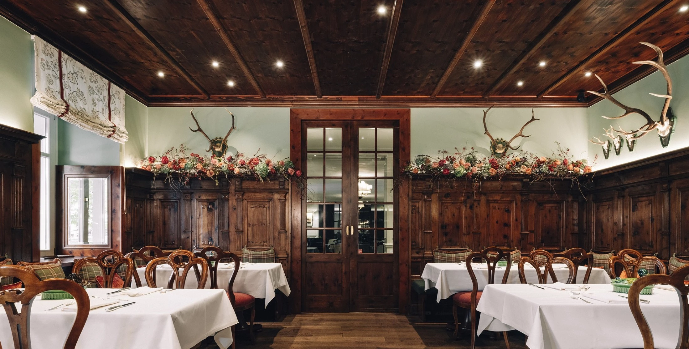 Rustikales Restaurant (Zirbelzimmer) mit dunklem Holzinterieur, weißen Tischdecken, Hirschgeweihen an der Wand und karierten Polsterbänken.