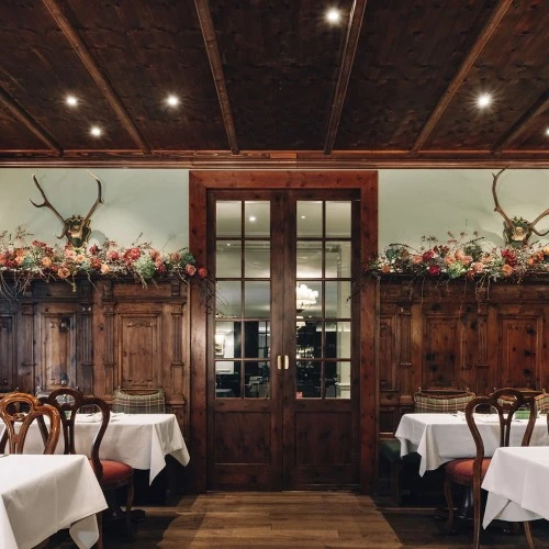 Rustikales Restaurant (Zirbelzimmer) mit dunklem Holzinterieur, weißen Tischdecken, Hirschgeweihen an der Wand und karierten Polsterbänken.