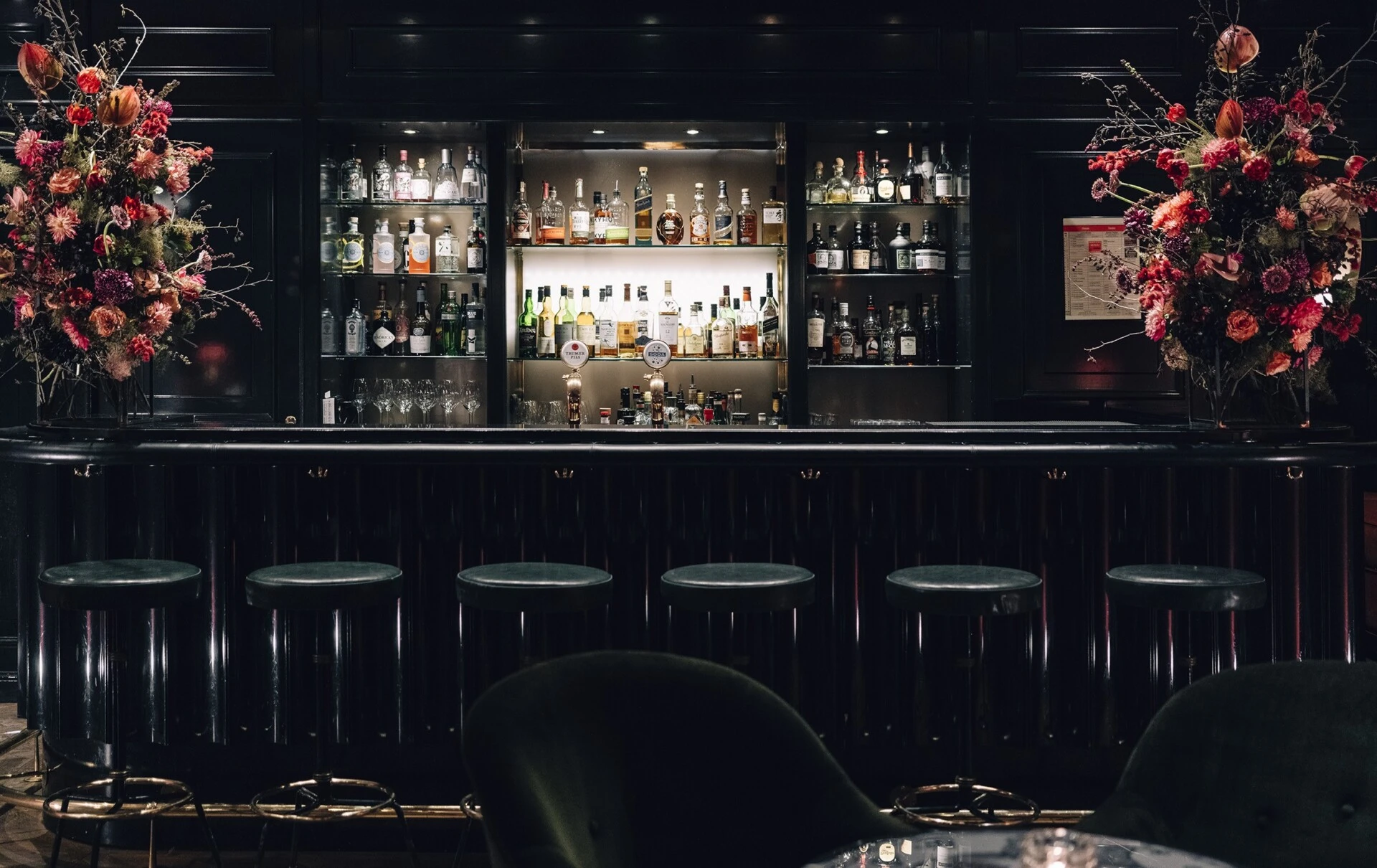 Stilvolle Hotelbar mit eleganter schwarzer Theke und sechs gepolsterten Barhockern. Hinter der Bar befindet sich ein beleuchtetes Regal mit einer großen Auswahl an Spirituosen und Gläsern