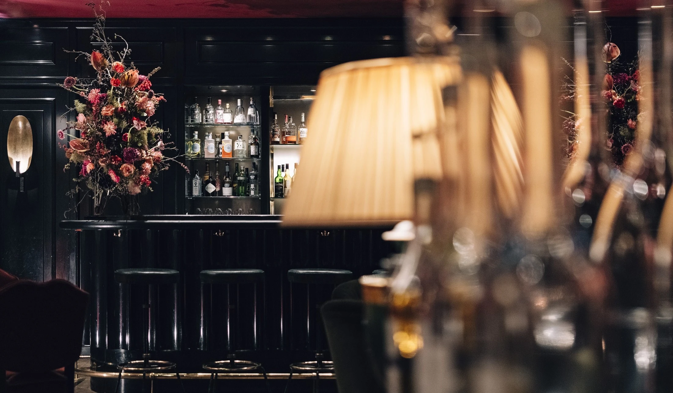 Elegante Bar mit dunkler Theke und stilvollen Barhockern. Im Hintergrund stehen zahlreiche hochwertige Spirituosen in einem beleuchteten Regal. Auf der Theke sorgt ein großes, farbenfrohes Blumenarrangement für einen edlen Akzent.