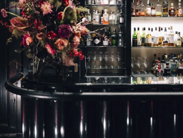 Eine elegante Bar mit dunkler, glänzender Theke und zwei runden Hockern im Vordergrund. Dahinter ein gut bestücktes Regal mit hochwertigen Spirituosen, Gläsern und stilvoller Beleuchtung. Links davon steht ein opulenter, farbenfroher Blumenstrauß, der dem Raum eine luxuriöse Note verleiht. Die Atmosphäre wirkt exklusiv und einladend – perfekt für einen stilvollen Drink.