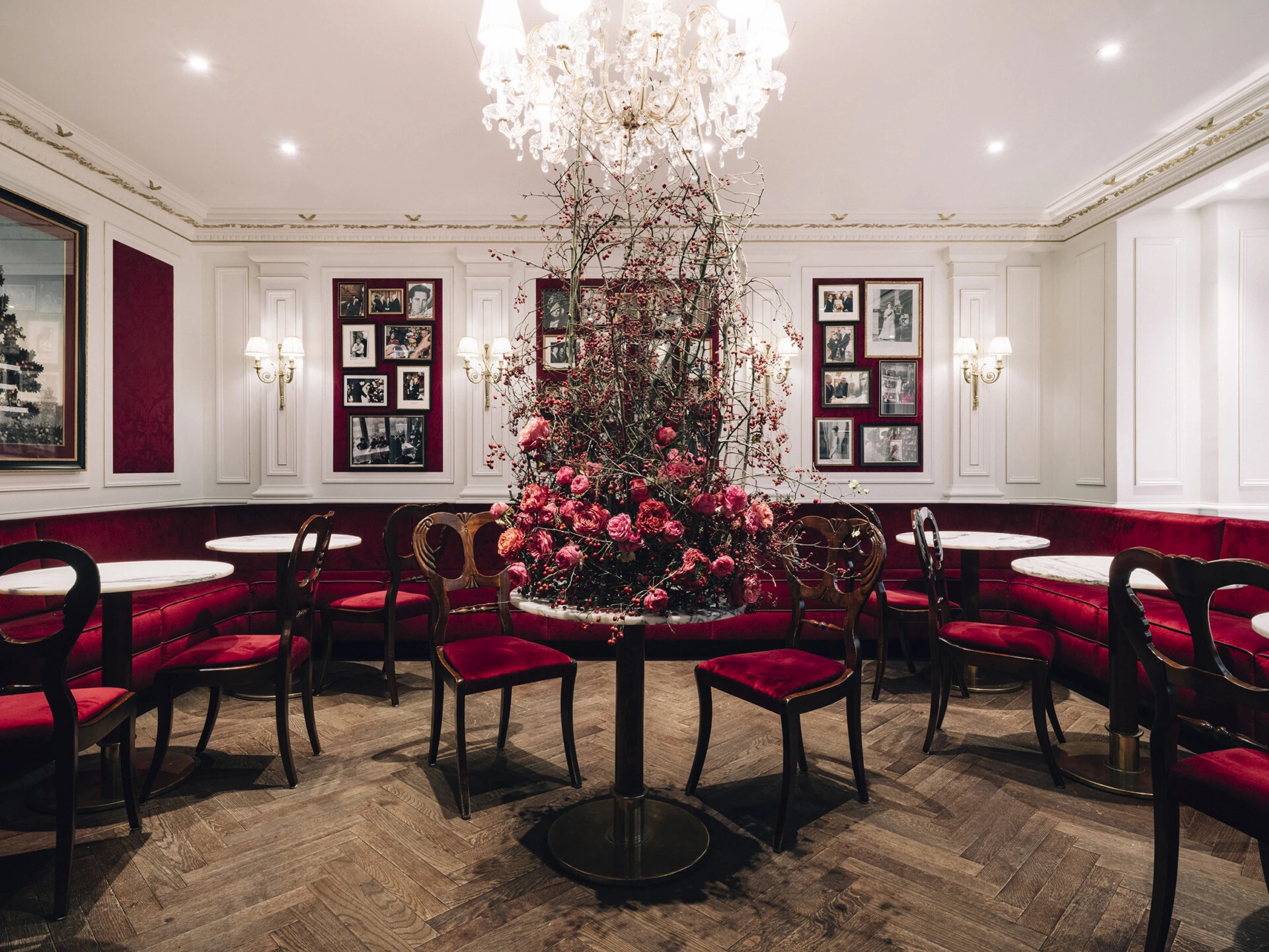 Ein stilvolles Kaffeehaus mit eleganten Marmortischen und samtroten Sitzbänken. Im Zentrum steht ein kunstvoll arrangiertes Blumenbouquet in Rosa- und Rottönen. Der Raum ist mit goldenen Wandlampen, weißen Wandvertäfelungen und gerahmten Fotografien dekoriert. Ein großer Kronleuchter rundet das edle Ambiente ab.