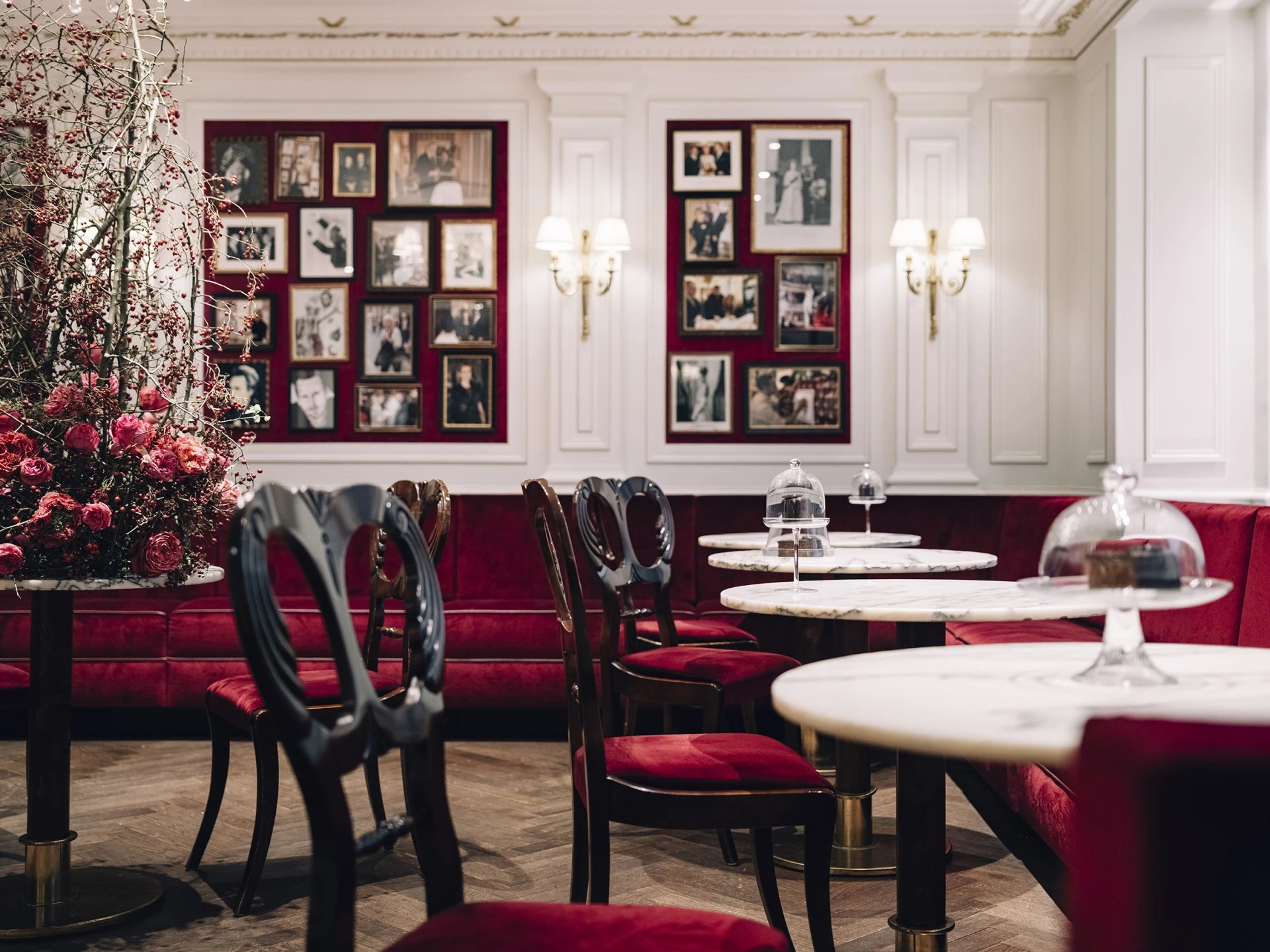 Ein eleganter Salon mit weißen Wandpaneelen, samtroten Polstermöbeln und Marmortischen. Auf den Tischen stehen unter Glasglocken präsentierte Stücke der Original Sacher-Torte. An den Wänden hängen zahlreiche gerahmte Fotos prominenter Gäste. Links ein opulentes Blumenarrangement, das dem Raum eine festliche Note verleiht.