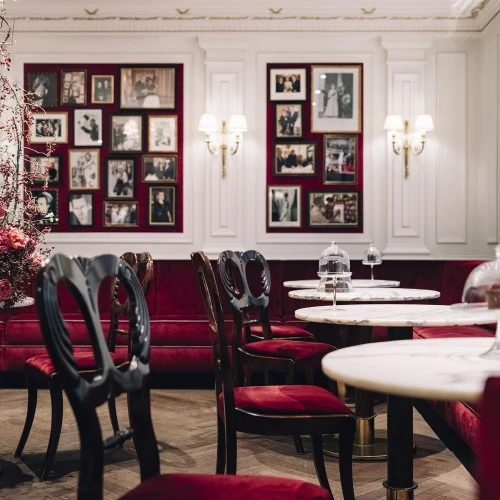 Ein eleganter Salon mit weißen Wandpaneelen, samtroten Polstermöbeln und Marmortischen. Auf den Tischen stehen unter Glasglocken präsentierte Stücke der Original Sacher-Torte. An den Wänden hängen zahlreiche gerahmte Fotos prominenter Gäste. Links ein opulentes Blumenarrangement, das dem Raum eine festliche Note verleiht.